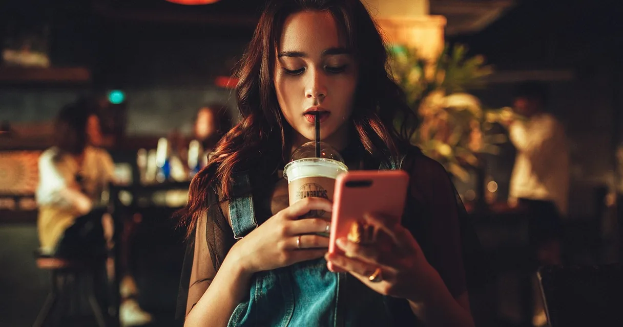 Mulher com copo de café olhando o celular em um bar com iluminação baixa